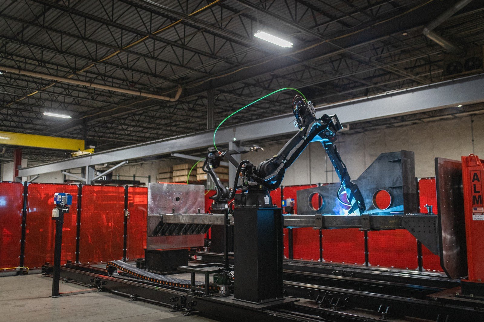 A robotic welding system welds a large, irregular structural steel weldment inside a fabrication facility.